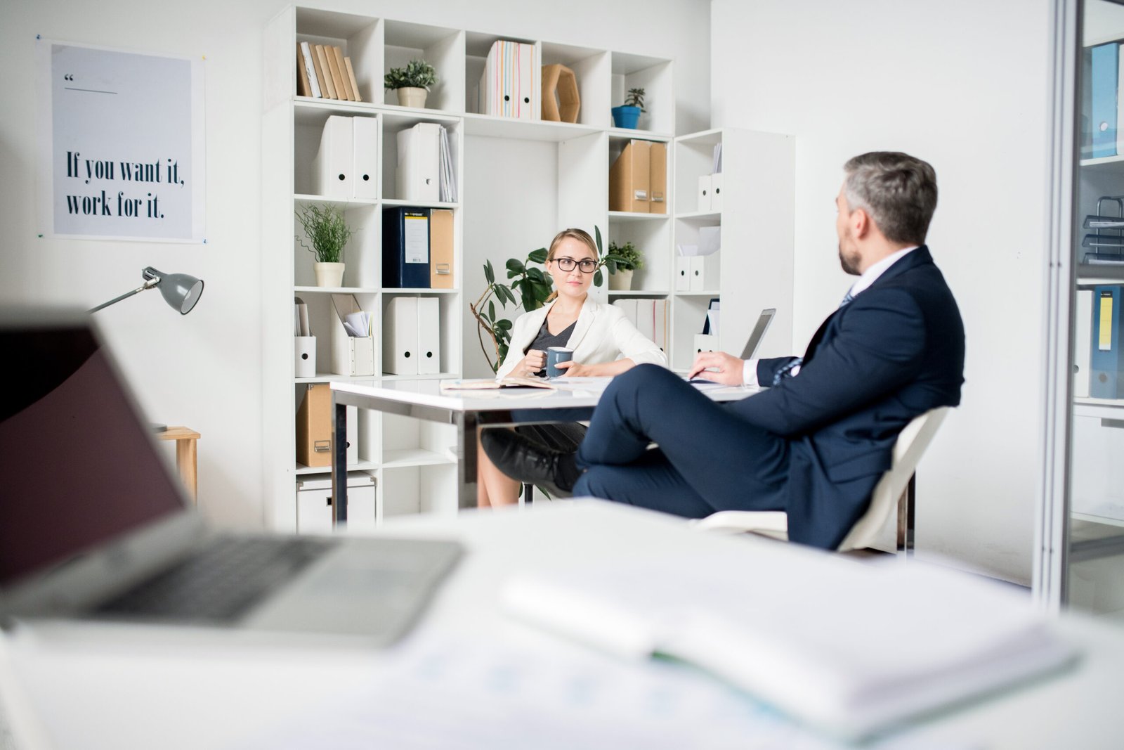 Confident business executives in formalwear sitting at table in modern office with bookshelf and motivated banner and discussing project plan while considering their next steps